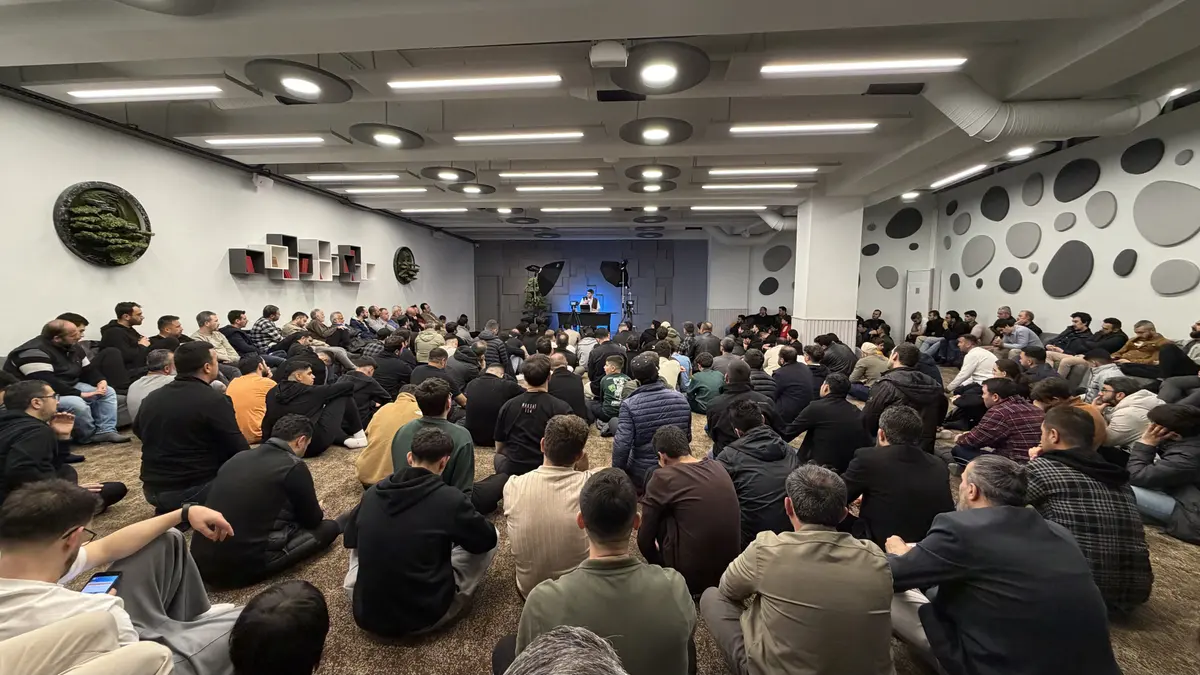 Maksat 114 ders salonu: yere oturmuş kalabalık bir topluluk, önde mavi ışıklı sahnede konuşmacı